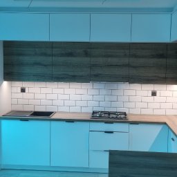 Tomasz Kuczynski - Nowoczesna kuchnia z białymi frontami i drewnopodobnymi szafkami górnymi, oświetleniem podszafkowym i białymi płytkami metra na ścianie, z piekarnikiem w zabudowie i płytą gazową.