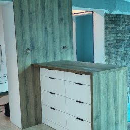 Tomasz Kuczynski - Wyspa kuchenna z białymi frontami szuflad i blatem w kolorze drewna, z widocznymi przygotowaniami do instalacji elektrycznej w ścianie z okładziną drewnopodobną, na błyszczącej podłodze w trakcie...