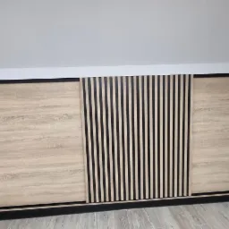 Szafa wnękowa z przesuwnymi drzwiami w kolorze jasnego drewna z czarnymi akcentami, dopasowana do skosu poddasza.