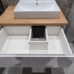 Nowoczesna łazienka z białą umywalką nablatową na blacie z drewna oraz szafką z szufladą z połyskiem, widoczny fragment toalety wiszącej.