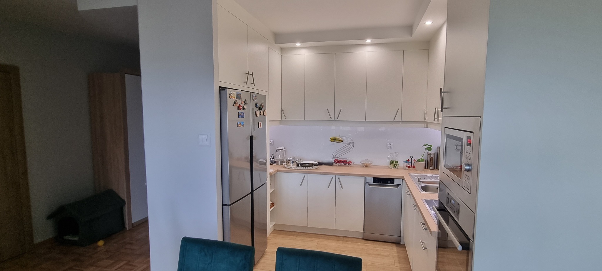 Jasna kuchnia na wymiar z białymi frontami, srebrną lodówką z magnesami i drewnianym blatem, widok z jadalni.