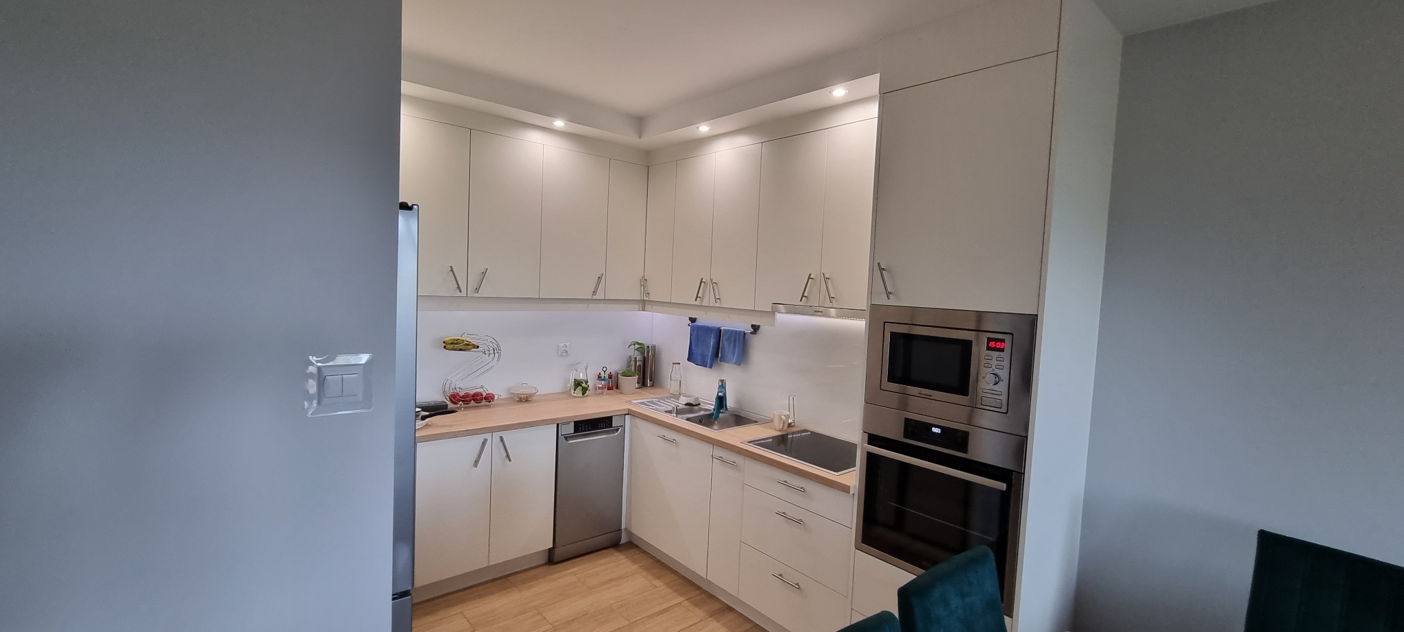 Jasna kuchnia z białymi szafkami, drewnianym blatem, wbudowanym piekarnikiem i mikrofalówką, widziana z perspektywy narożnej