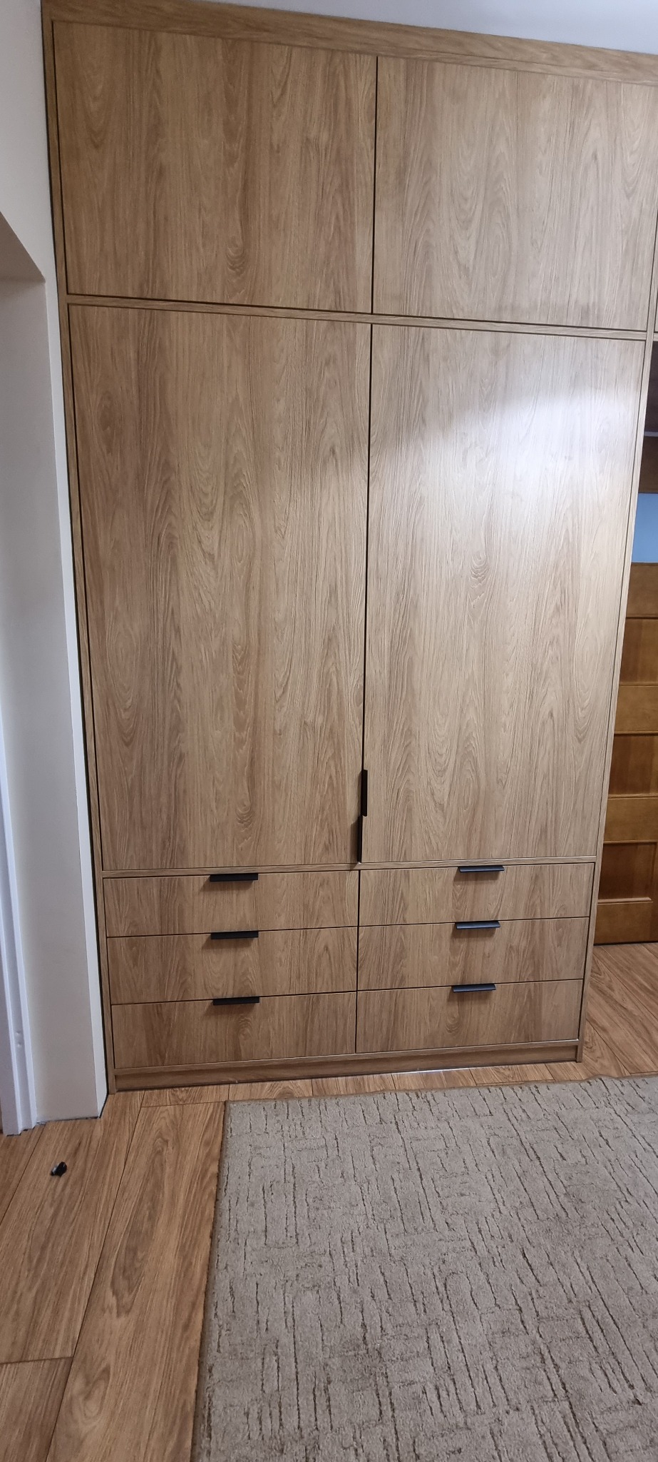 Wysoka szafa wnękowa z frontami imitującymi jasne drewno, z czarnymi, minimalistycznymi uchwytami szuflad, na tle parkietu i dywanu.