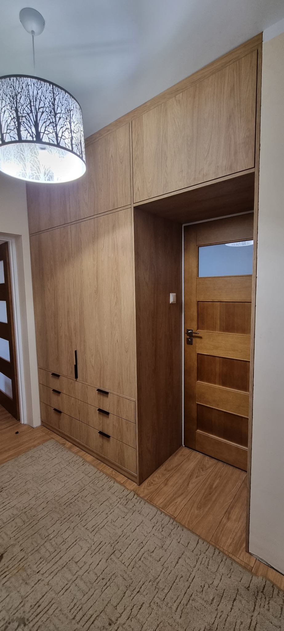 Drewniana szafa wnękowa z szufladami i czarnymi uchwytami, obok drewnianych drzwi z szybą, oświetlona lampą z motywem drzew.