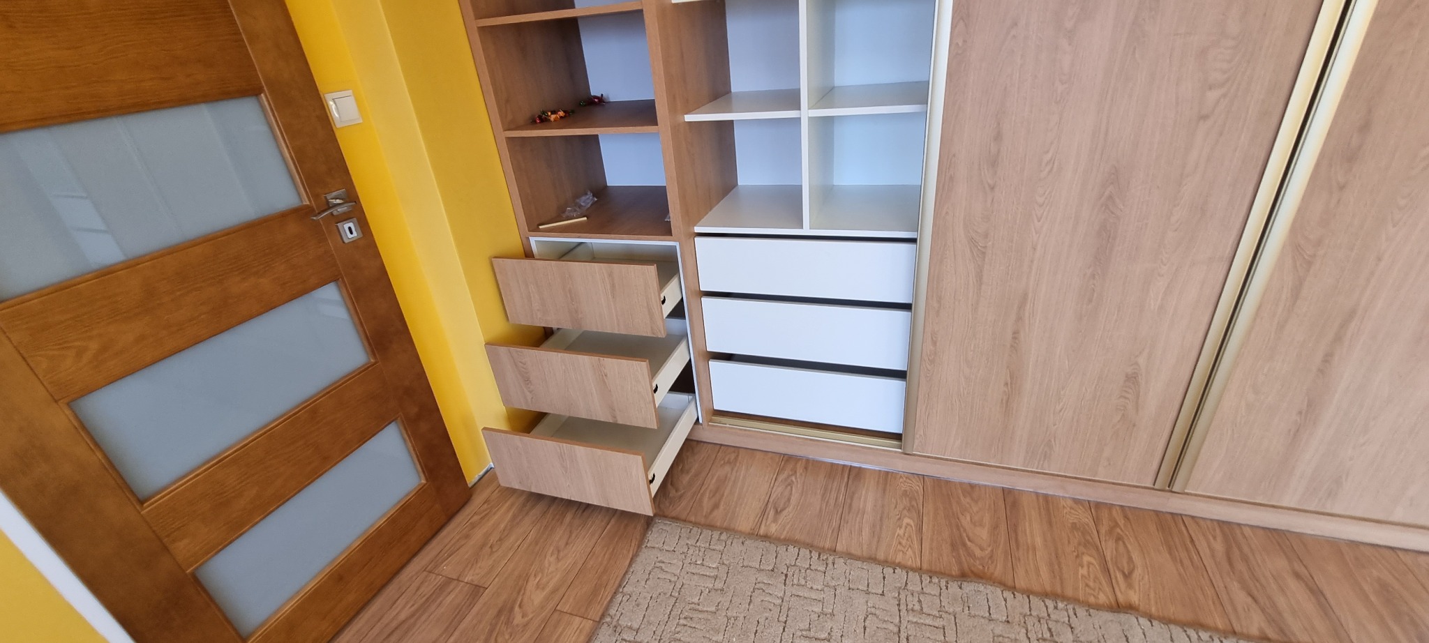 Wnętrze szafy wnękowej z otwartymi szufladami o drewnianym wykończeniu, widoczna drewniana podłoga i drzwi z szybami.