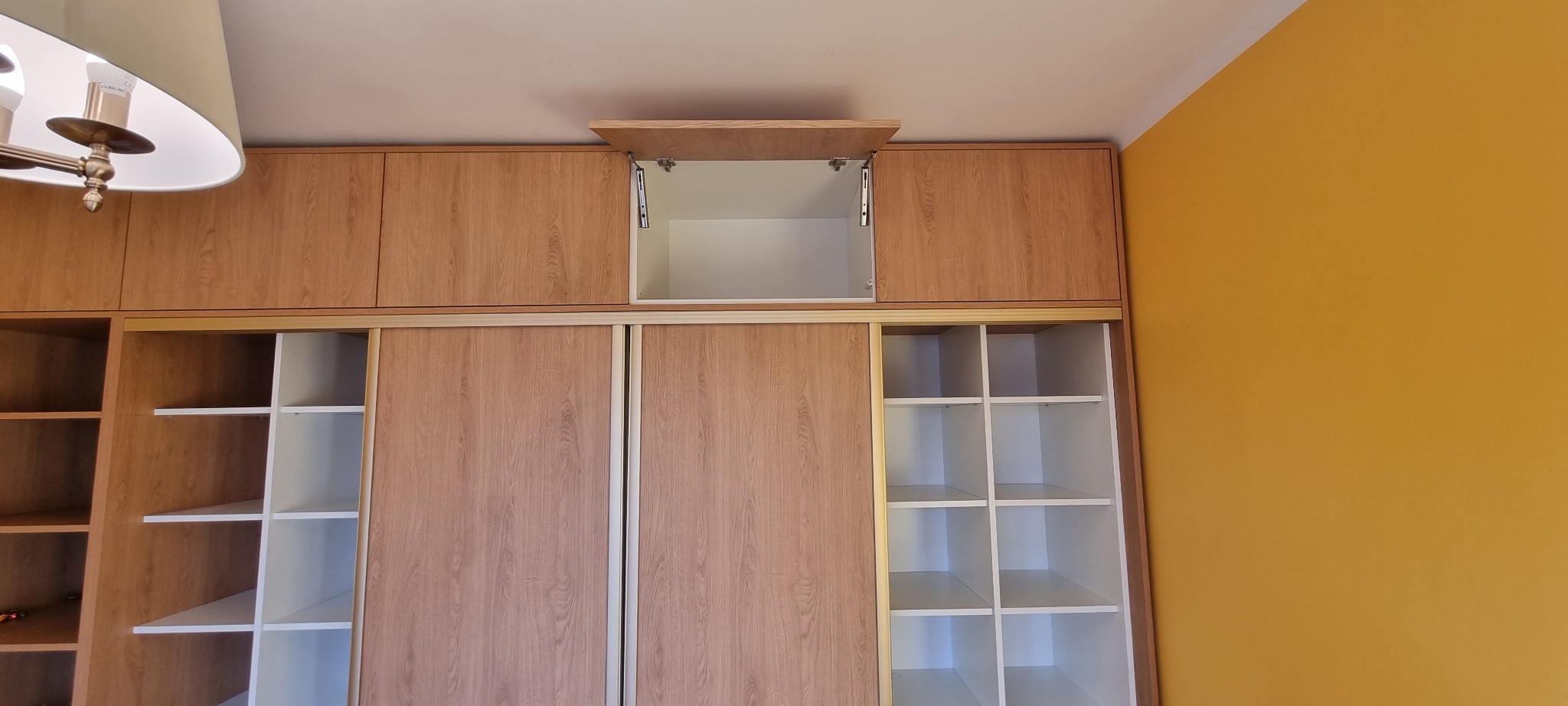 Wysoka szafa wnękowa z przesuwnymi drzwiami o jasnym, drewnianym wykończeniu, z otwartą szafką górną na zawiasach i widocznymi półkami po bokach.