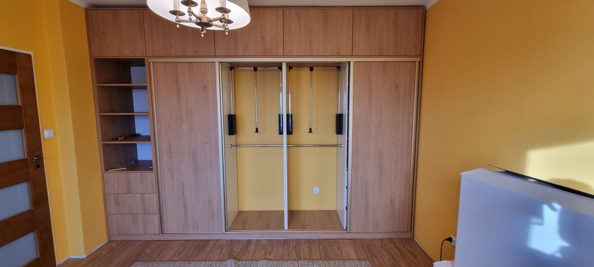 Szafa wnękowa z przesuwnymi drzwiami i systemem pantografów w Ostrowi Mazowieckiej, z otwartą częścią regałową po lewej stronie, na tle żółtej ściany.