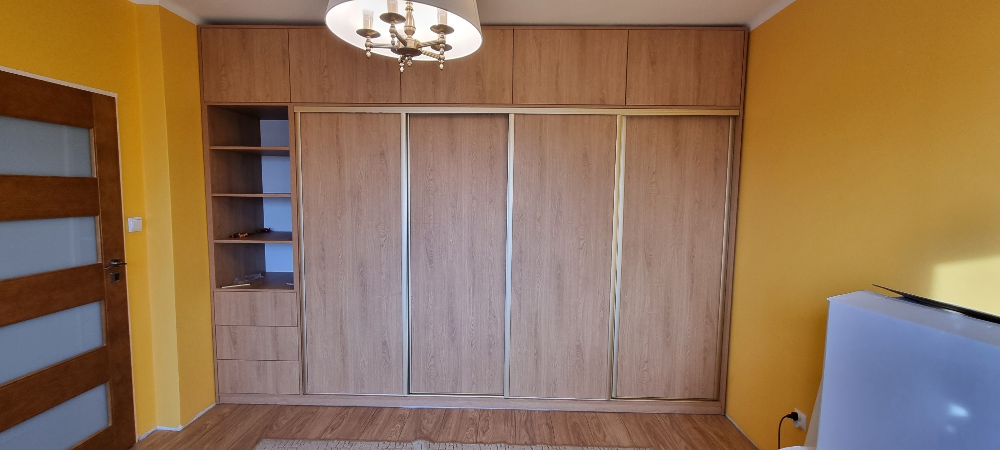Wnękowa szafa z przesuwnymi drzwiami w odcieniach dębu, z otwartym regałem po lewej stronie, uzupełniona o szafki górne, w pokoju z żółtymi ścianami.