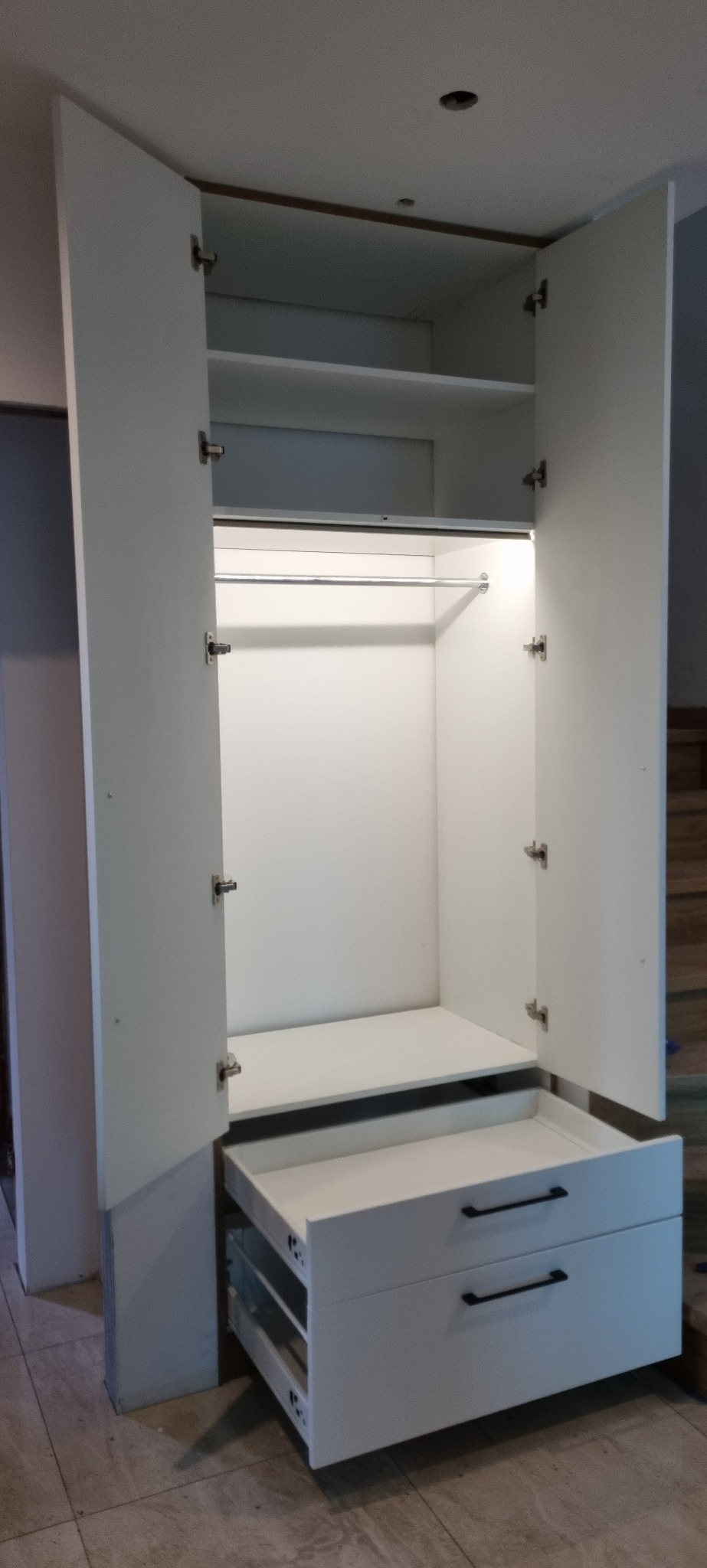 Wnętrze białej szafy wnękowej z otwartymi drzwiami, widoczny drążek na ubrania, półki i dwie szuflady z czarnymi uchwytami.