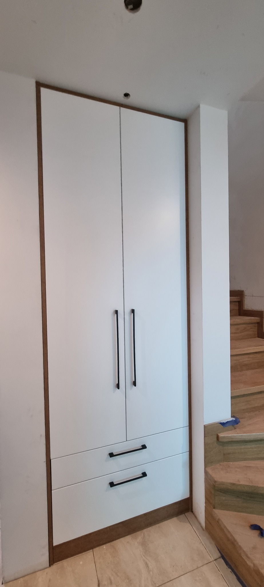 Biała szafa wnękowa z dwoma szufladami i czarnymi uchwytami, obudowana drewnianą ramą, widoczna obok schodów.