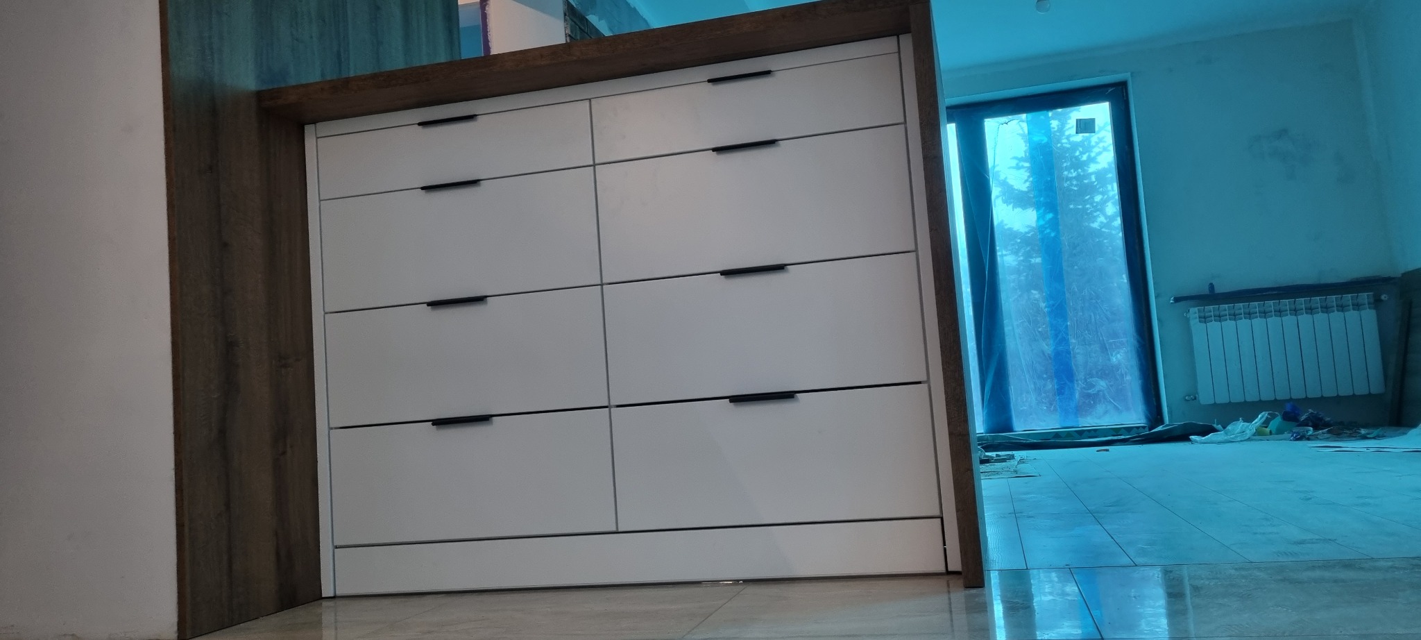 Wbudowana szafa z białymi frontami szuflad i ciemnobrązową ramą, widoczna podczas montażu w pokoju z niebieską folią ochronną na oknie i grzejnikiem.