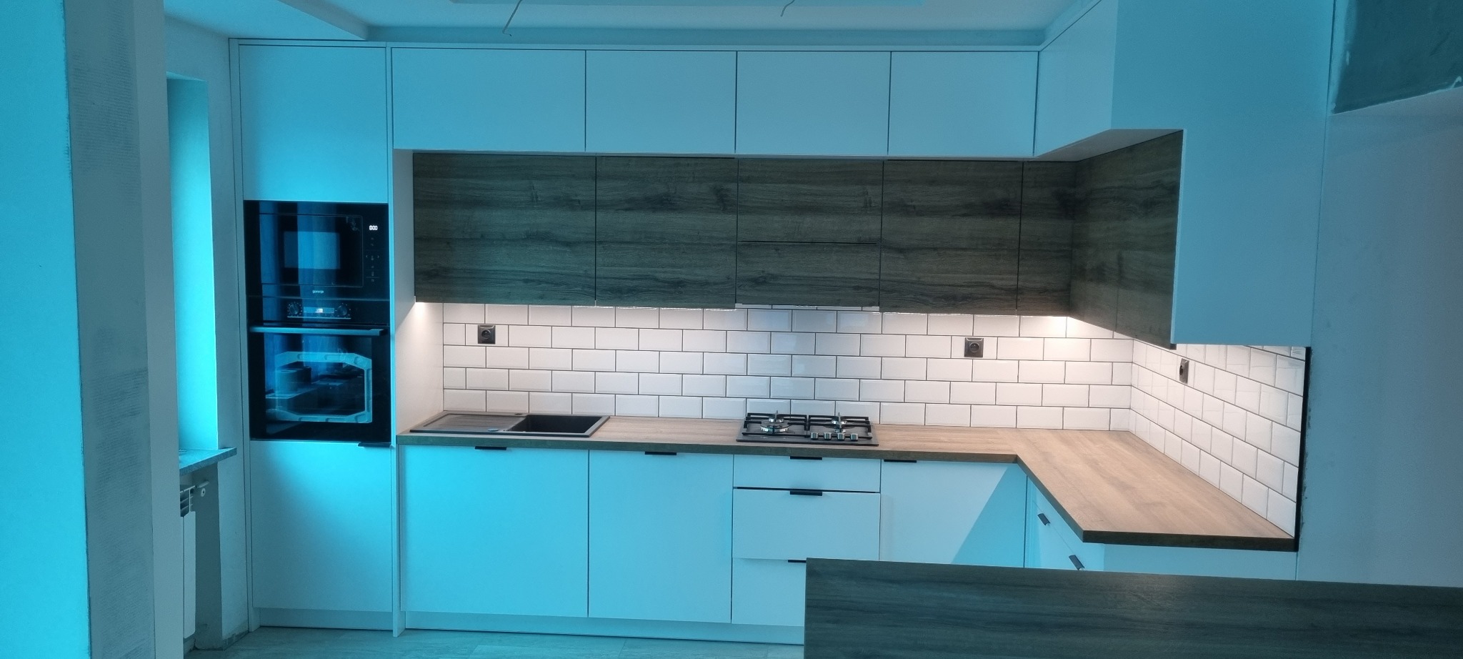 Nowoczesna kuchnia z białymi frontami i drewnopodobnymi szafkami górnymi, oświetleniem podszafkowym i białymi płytkami metra na ścianie, z piekarnikiem w zabudowie i płytą gazową.