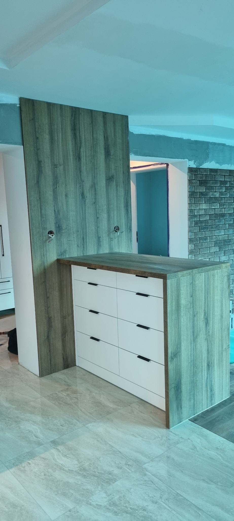 Wyspa kuchenna z białymi frontami szuflad i blatem w kolorze drewna, z widocznymi przygotowaniami do instalacji elektrycznej w ścianie z okładziną drewnopodobną, na błyszczącej podłodze w trakcie...