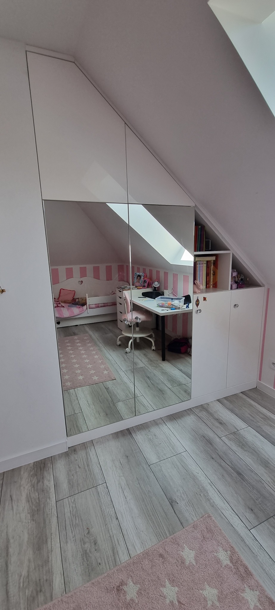 Biała szafa wnękowa z lustrzanymi drzwiami w pokoju dziecięcym na poddaszu, odbijająca łóżko i biurko.