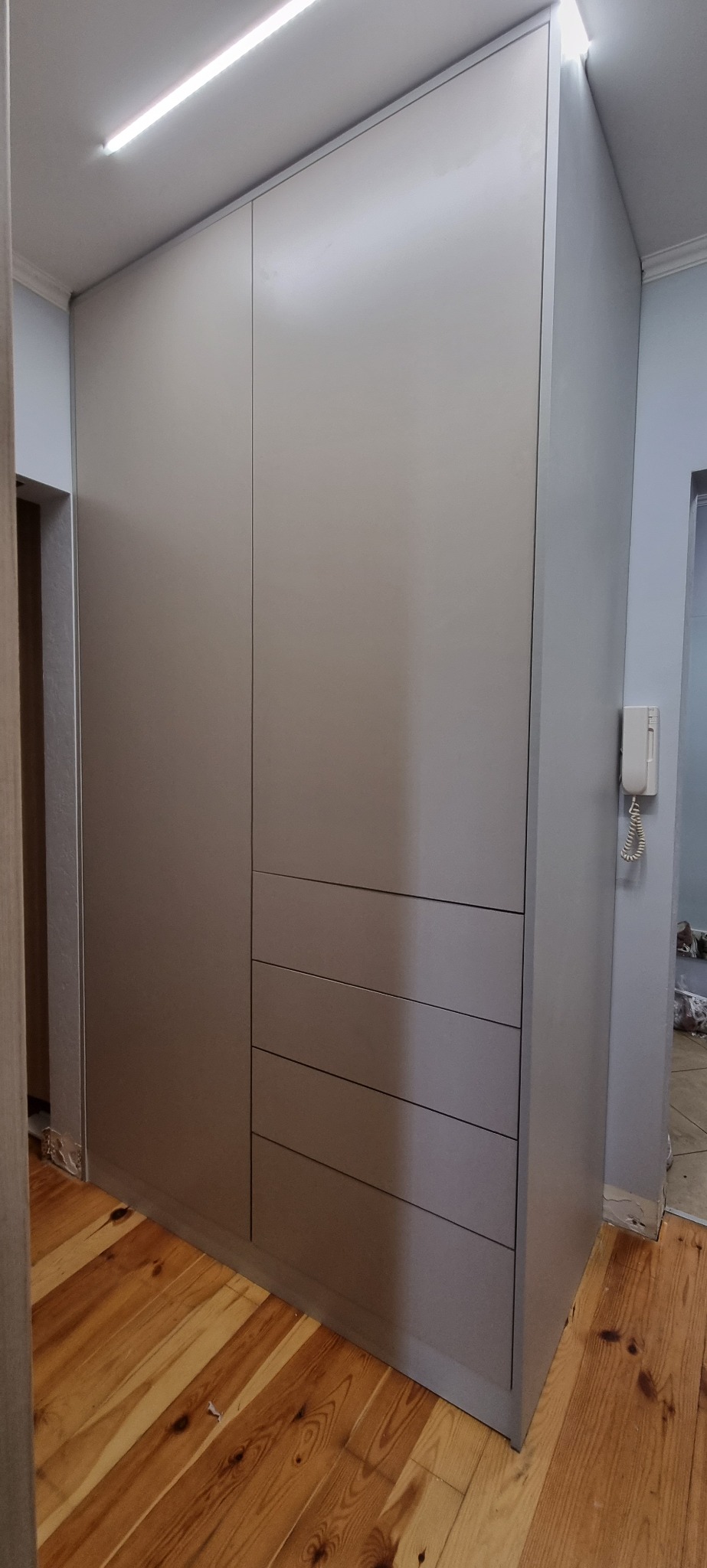 Szara szafa wnękowa z czterema szufladami, dopasowana do drewnianej podłogi, z widocznym oświetleniem sufitowym LED i telefonem stacjonarnym na ścianie.