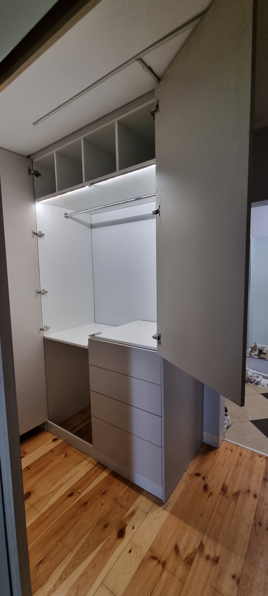 Wnętrze szafy wnękowej z półkami, drążkiem na ubrania i komodą z szufladami, oświetlone listwą LED, z otwartymi drzwiami i widoczną drewnianą podłogą.