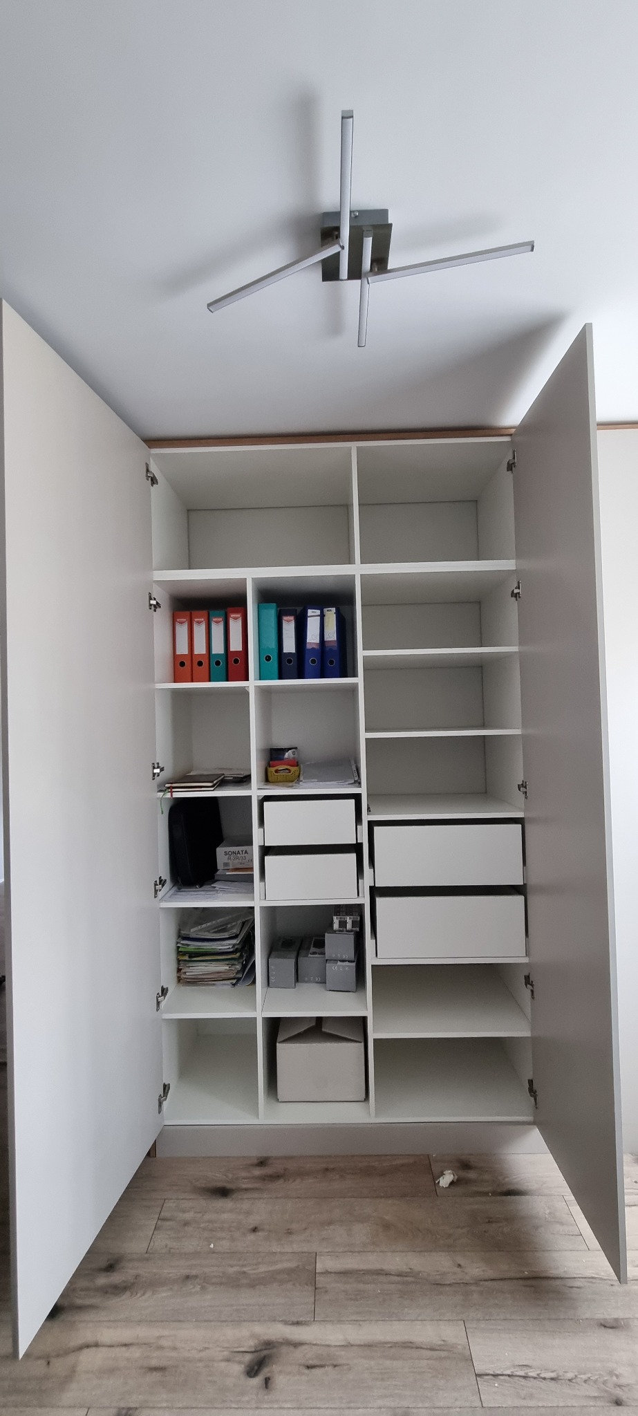 Wnętrze białej szafy wnękowej z otwartymi drzwiami, widoczne półki z segregatorami, dokumentami i pudełkami, oświetlenie sufitowe z metalowymi ramionami.