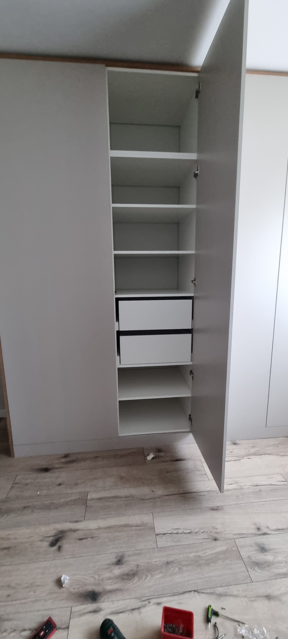 Biała szafa wnękowa z otwartymi drzwiami, widoczne półki i dwie szuflady, montaż szafy w trakcie prac wykończeniowych, na podłodze narzędzia.
