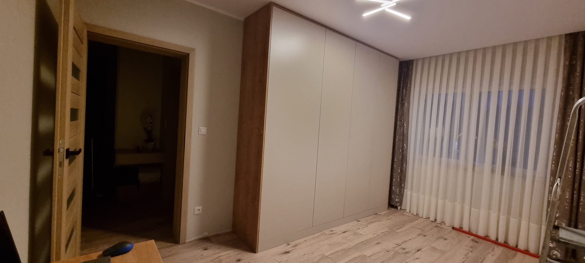 Duża, beżowa szafa wnękowa z drewnianymi bokami, dopasowana do drewnianej podłogi i ramy drzwi, z widoczną drabiną i zasłonami w tle.