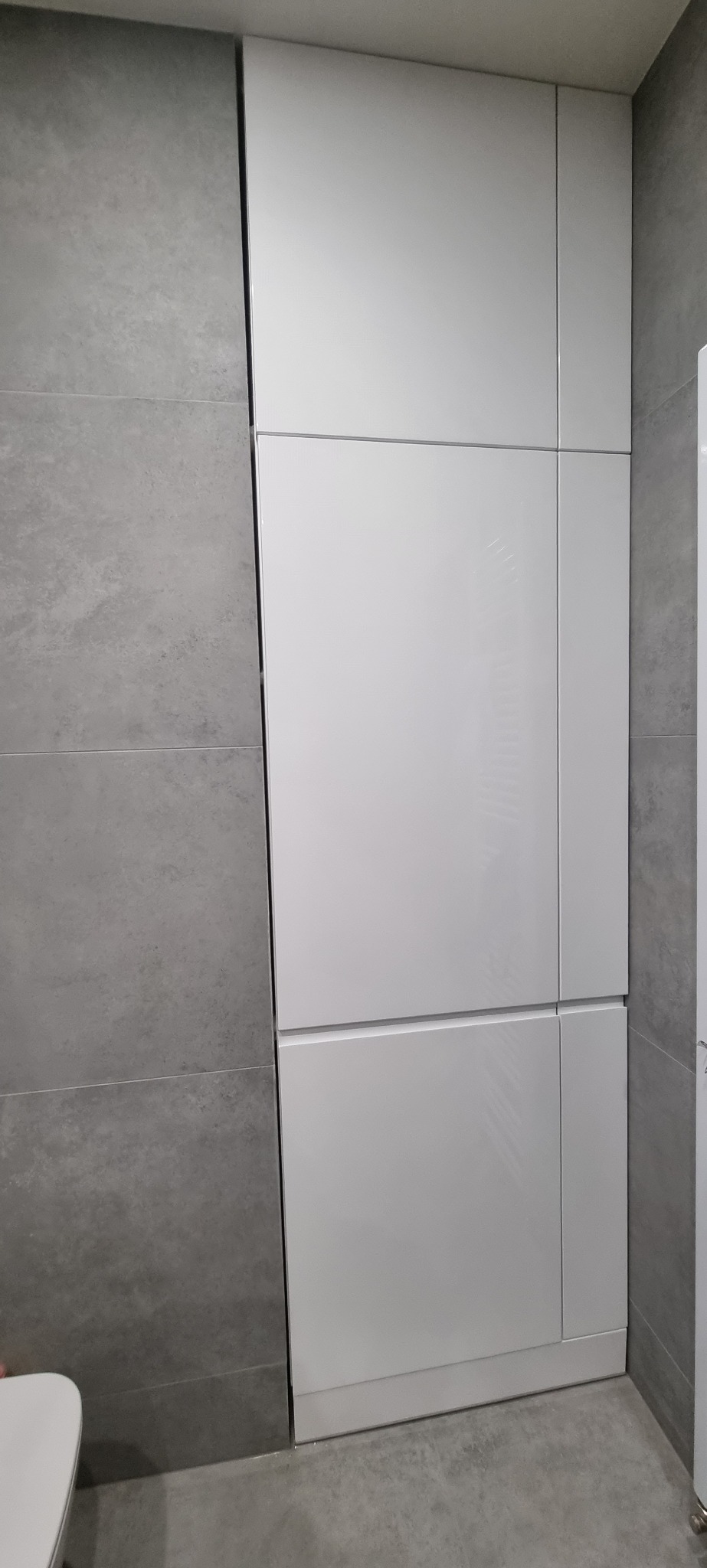 Biała szafa wnękowa z frontami na wysoki połysk, dopasowana do szarej ściany z płytek imitujących beton, widoczna z bliskiej perspektywy.