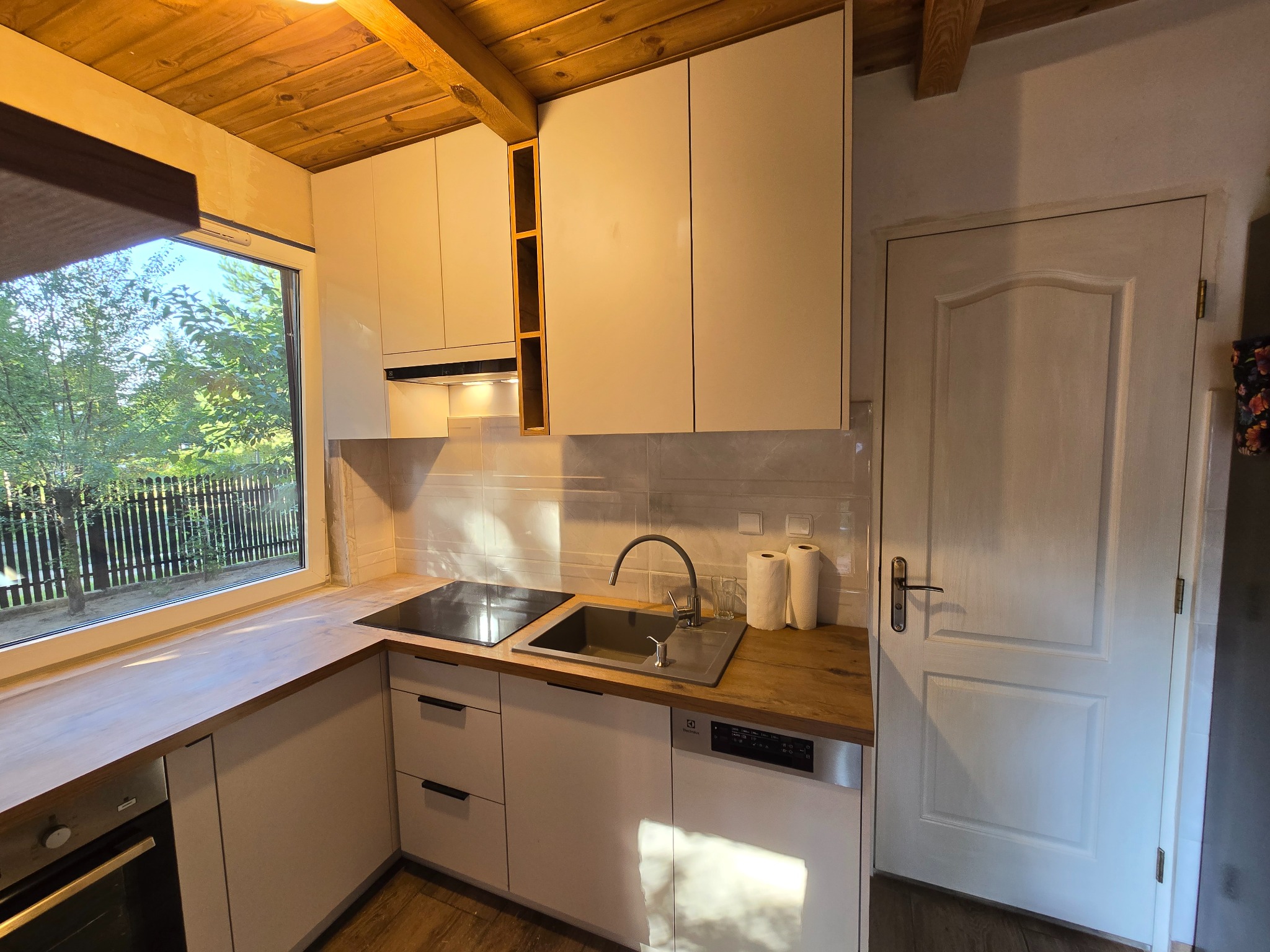 Jasna kuchnia z drewnianym blatem i widokiem na zieleń za oknem, drewniany sufit z belkami, białe fronty szafek z czarnymi uchwytami, zlewozmywak ze stali nierdzewnej.