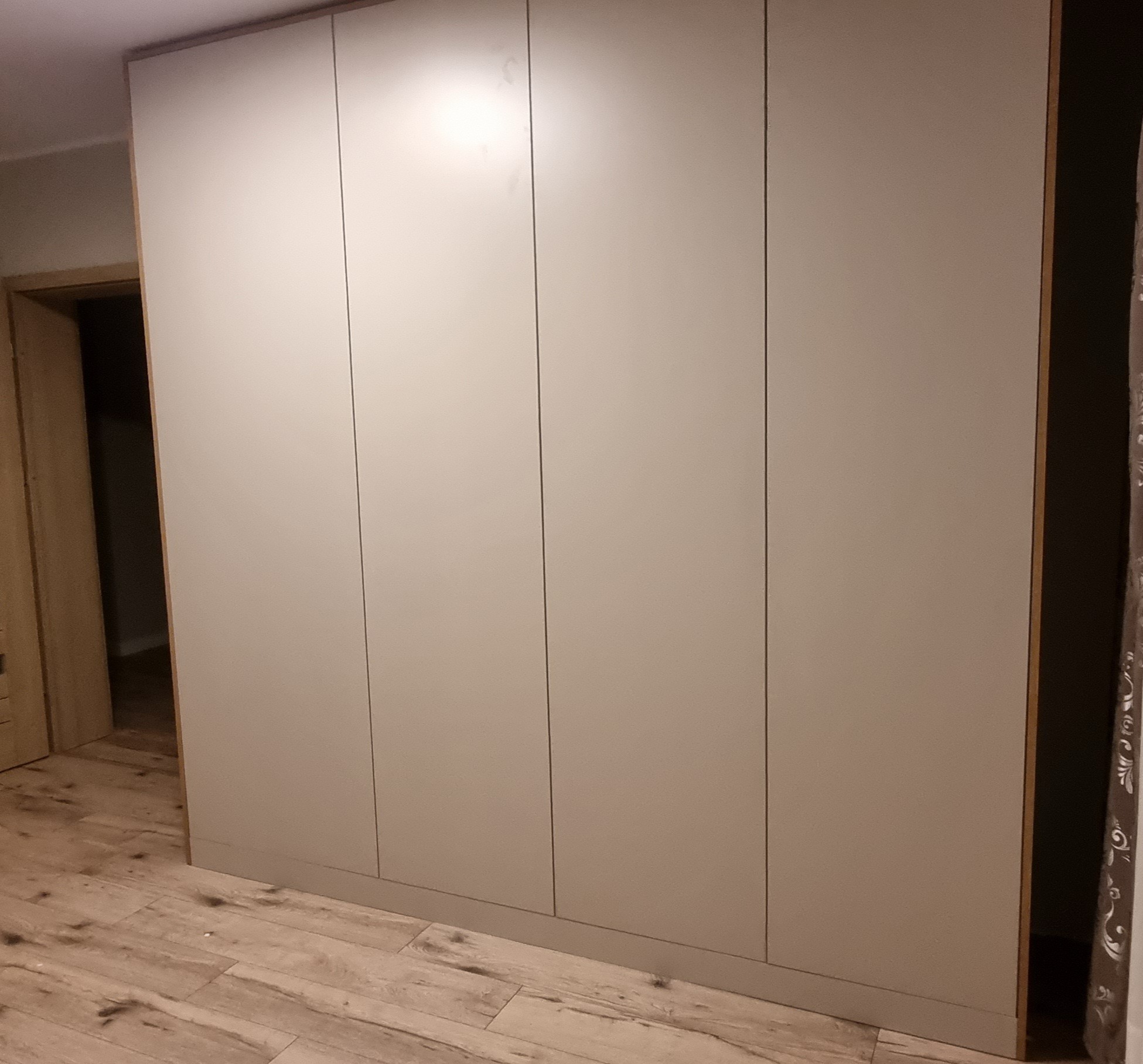 Czterodrzwiowa szafa wnękowa z gładkimi, beżowymi frontami, widoczna na tle drewnianej podłogi i częściowo otwartych drzwi do innego pomieszczenia.