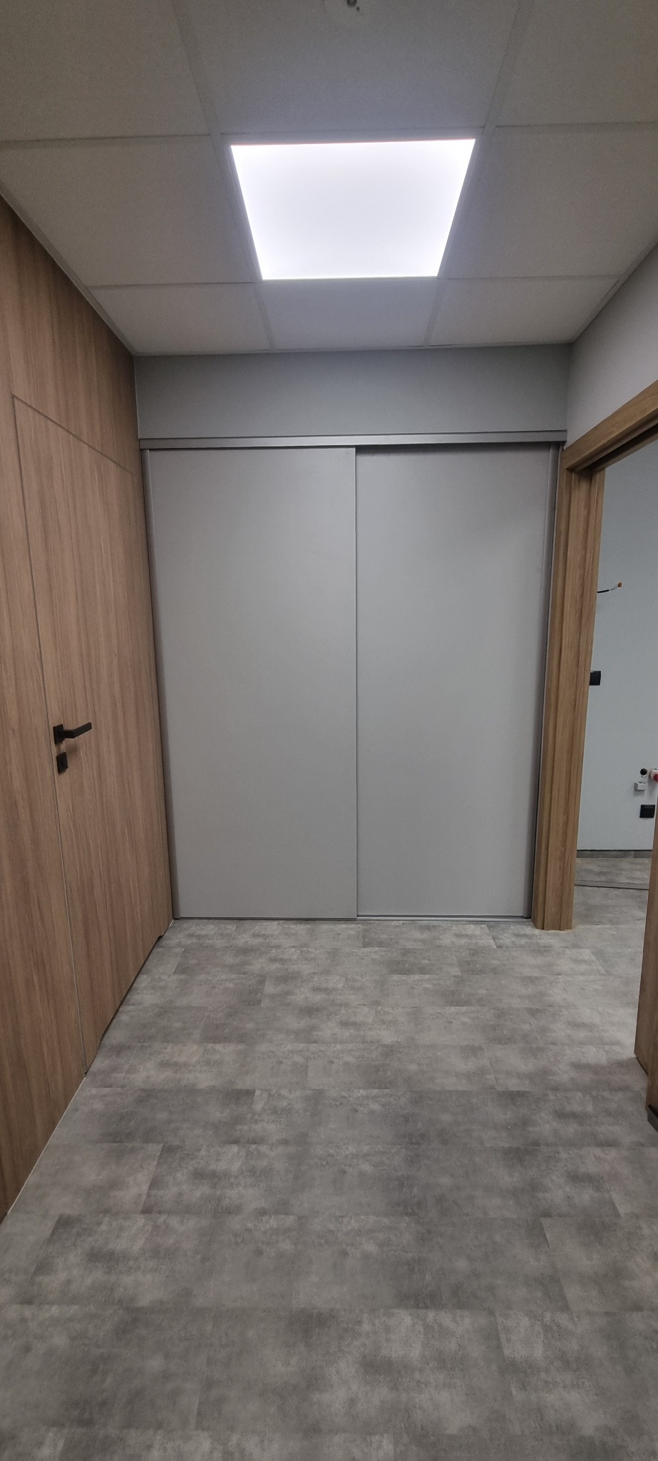 Szare, przesuwne drzwi szafy wnękowej z aluminiowymi prowadnicami, widok z perspektywy podłogi z szarymi panelami, drzwi zlicowane ze ścianą, obok drewniane drzwi wejściowe.