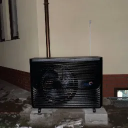 Czarna pompa ciepła marki FOX na betonowych fundamentach przed budynkiem w zimowej scenerii z resztkami śniegu na ziemi.