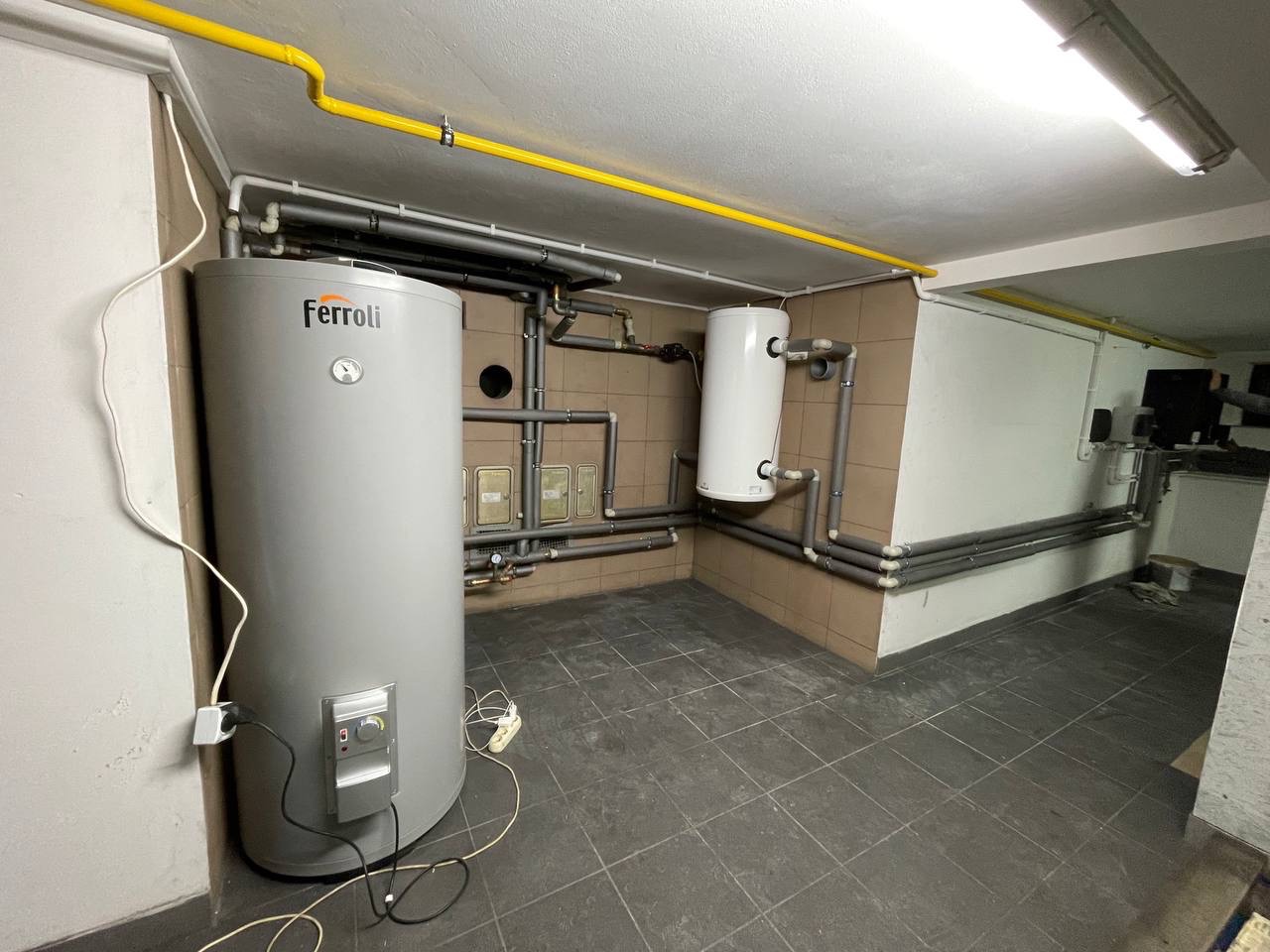 Szary, cylindryczny zbiornik Ferroli z widocznymi regulatorami i podłączeniami elektrycznymi, oraz mniejszy biały zbiornik, umieszczone w piwnicy z beżowymi płytkami i widocznymi rurami...