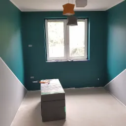 Poddasze w trakcie przygotowania do izolacji: widoczne płyty styropianowe, narzędzia, pomalowane ściany na zielono i szaro, okno z widokiem na drzewa.