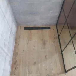 Wykończona kabina prysznicowa z odpływem liniowym w podłodze wyłożonej płytkami imitującymi drewno, szare płytki na ścianach i suficie, oraz czarna rama kabiny.