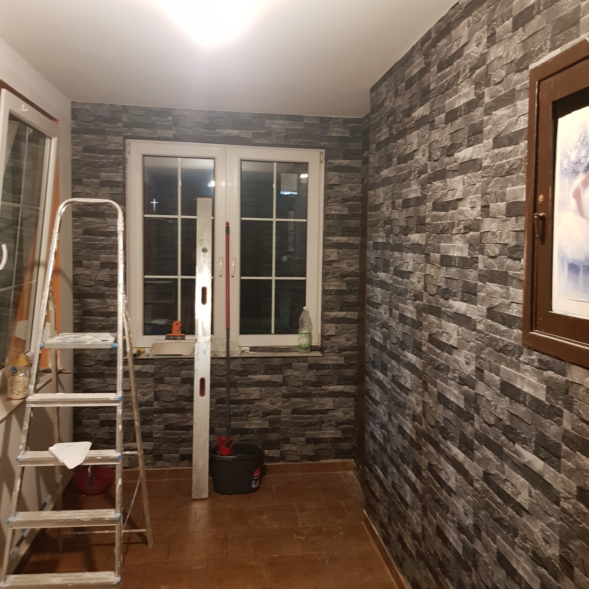 Wnętrze po wykończeniu ściany imitacją kamienia, widoczna drabina, poziomica, wiadro i okno.