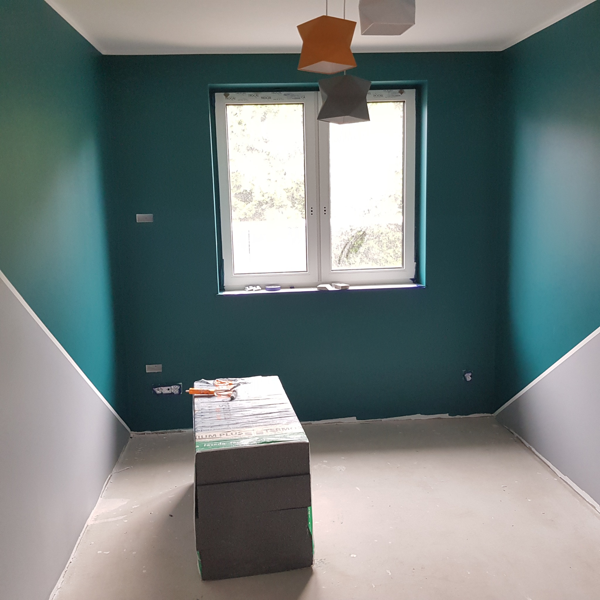 Poddasze w trakcie przygotowania do izolacji: widoczne płyty styropianowe, narzędzia, pomalowane ściany na zielono i szaro, okno z widokiem na drzewa.