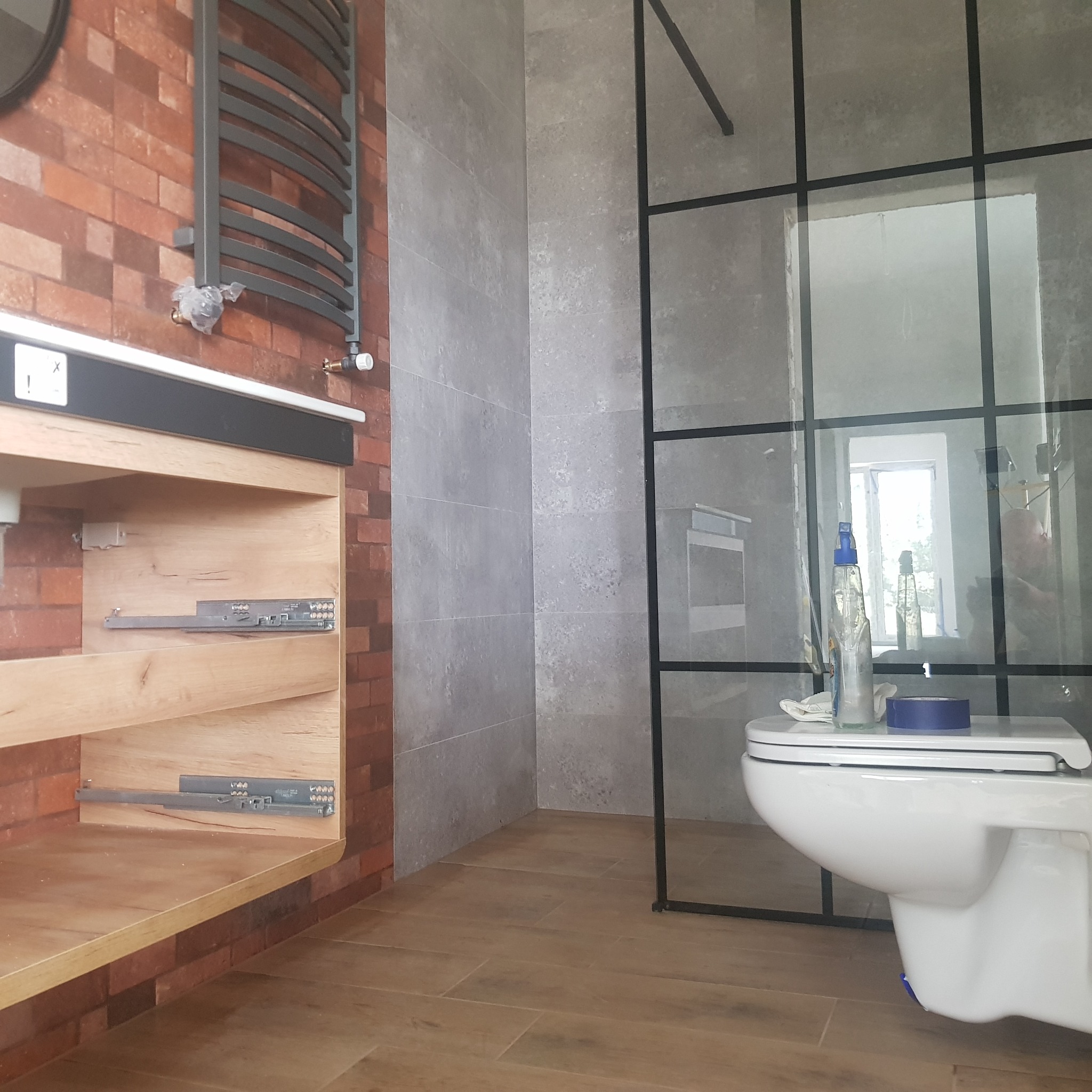 Nowoczesna łazienka w trakcie wykańczania: ściana z czerwonej cegły, grzejnik drabinkowy z folią ochronną, drewniana szafka pod umywalkę z wysuwanymi szufladami, szare płytki, kabina prysznicowa...