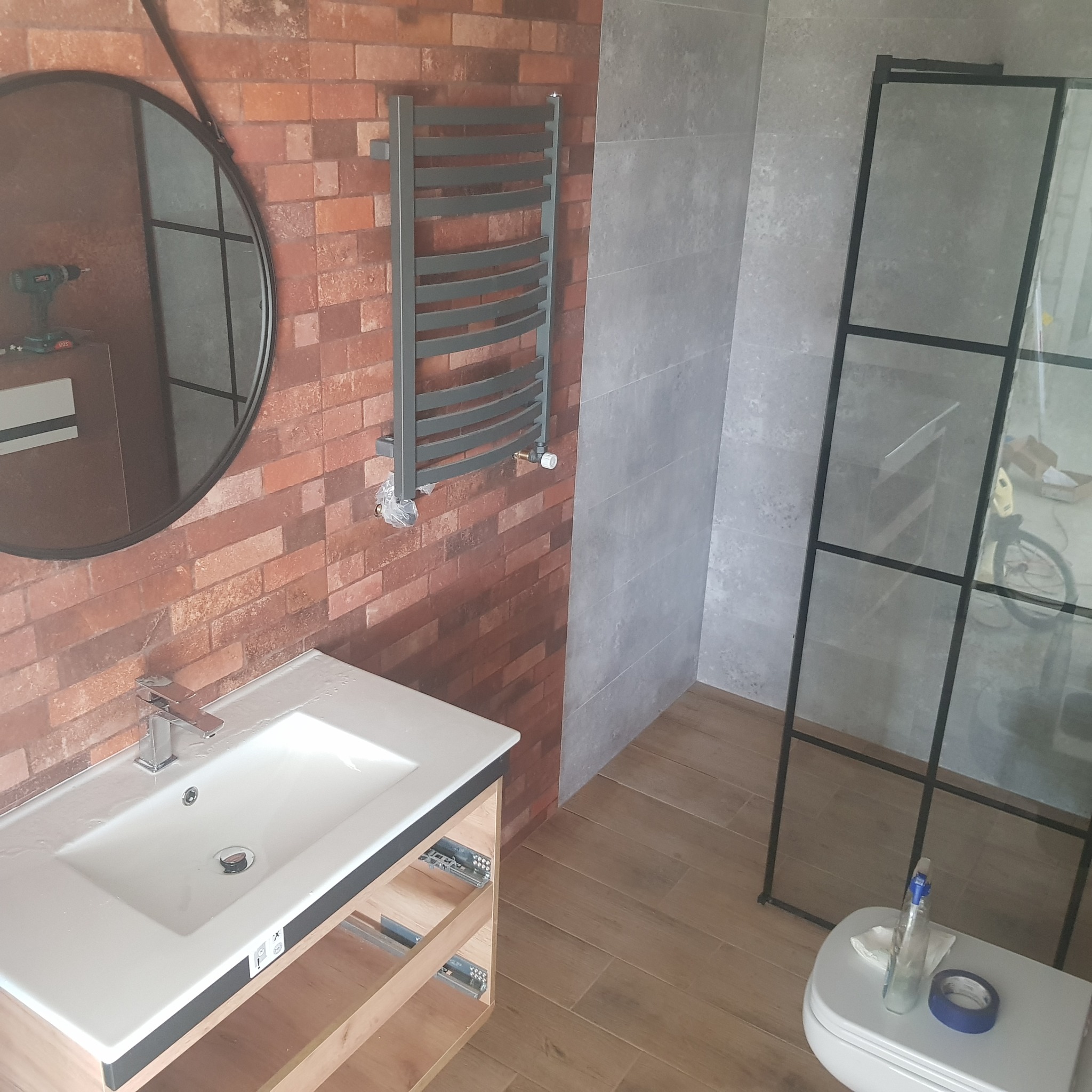 Łazienka w trakcie wykańczania: umywalka z szafką bez frontów, grzejnik drabinkowy, kabina prysznicowa z czarnymi profilami, ściana z cegły oraz narzędzia na sedesie.