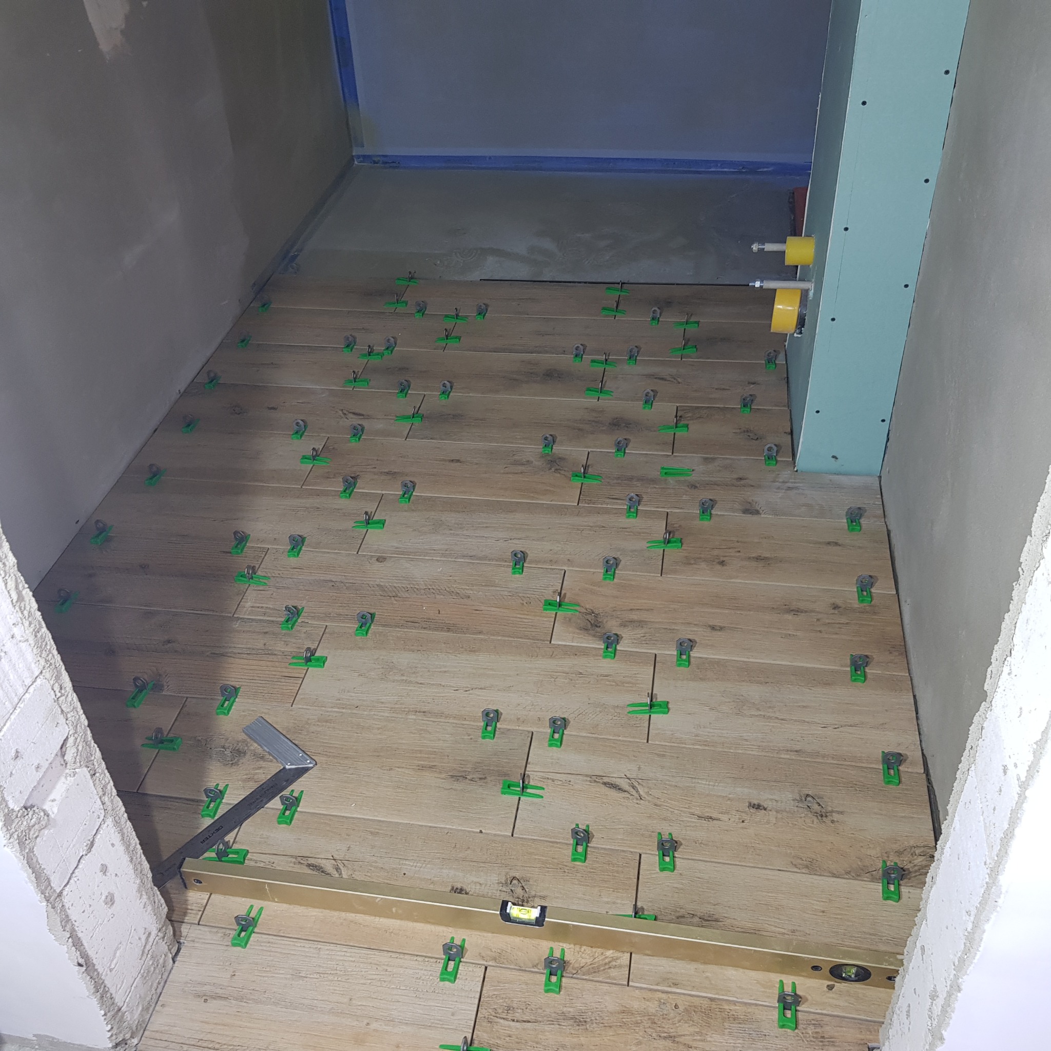 Montaż drewnopodobnych płytek podłogowych z użyciem poziomicy i klipsów poziomujących w Pucku, widoczne przygotowane ściany pod wykończenie.