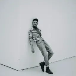 Mężczyzna w pasiastym swetrze i jeansach opiera się o białą ścianę, pozując do czarno-białego portretu.