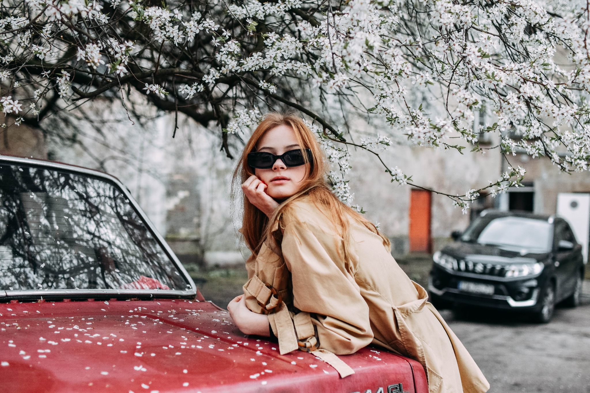 Młoda kobieta w okularach przeciwsłonecznych i beżowym płaszczu opiera się o czerwony samochód pokryty białymi płatkami, kwitnące drzewo w tle, drugi samochód w oddali.