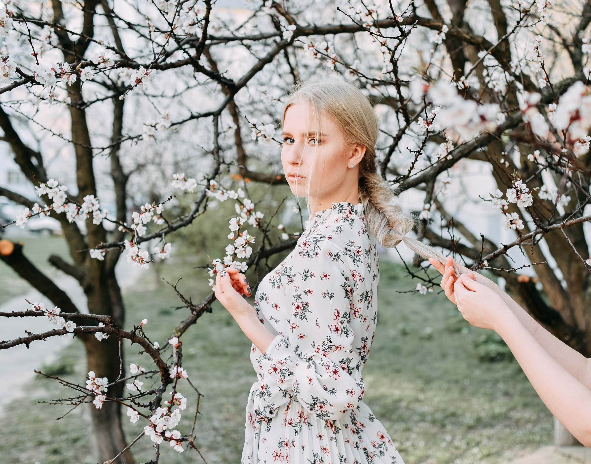 Kobieta w kwiecistej sukience pozuje na tle kwitnącej wiśni, druga osoba poprawia jej warkocz.