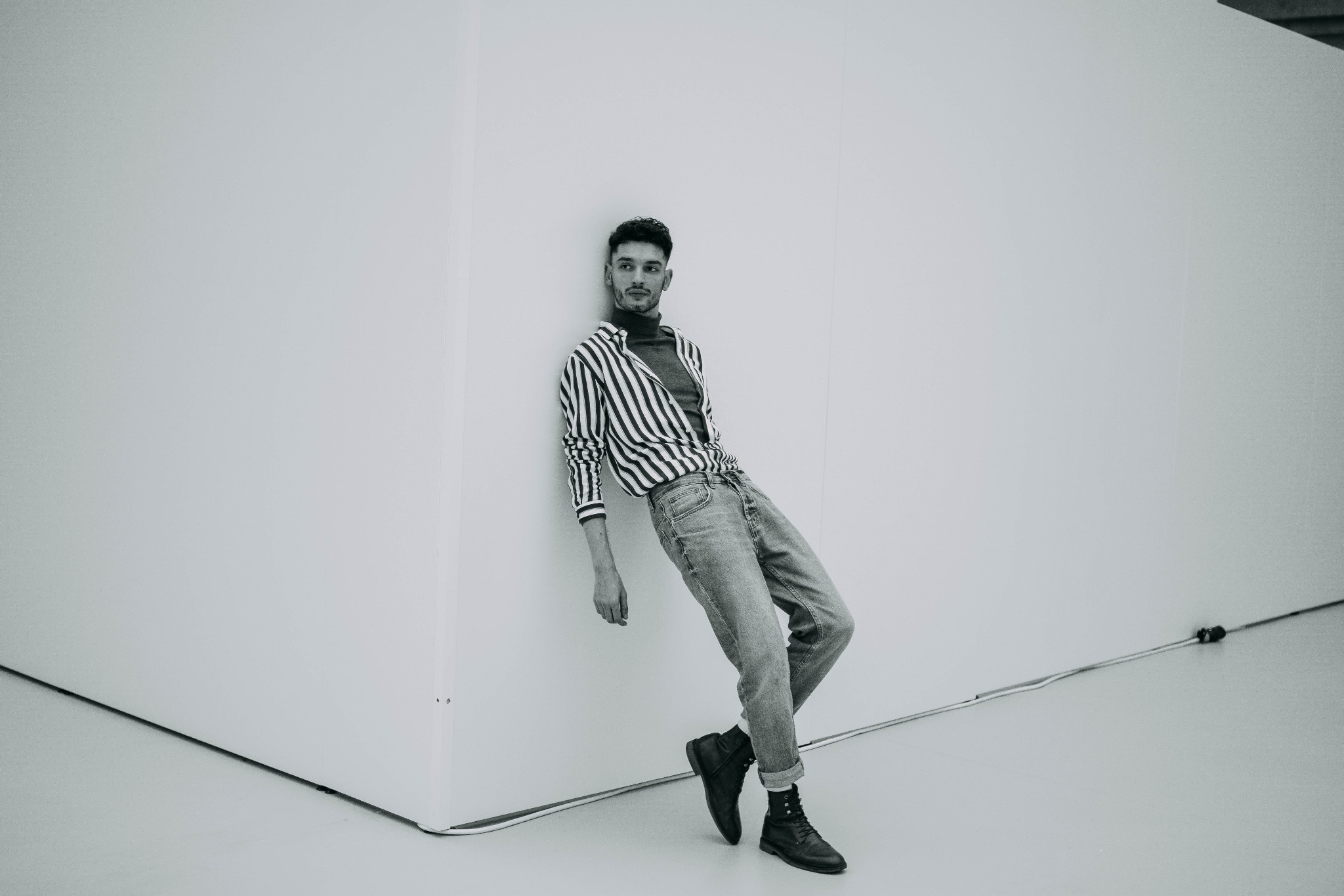 Mężczyzna w pasiastym swetrze i jeansach opiera się o białą ścianę, pozując do czarno-białego portretu.