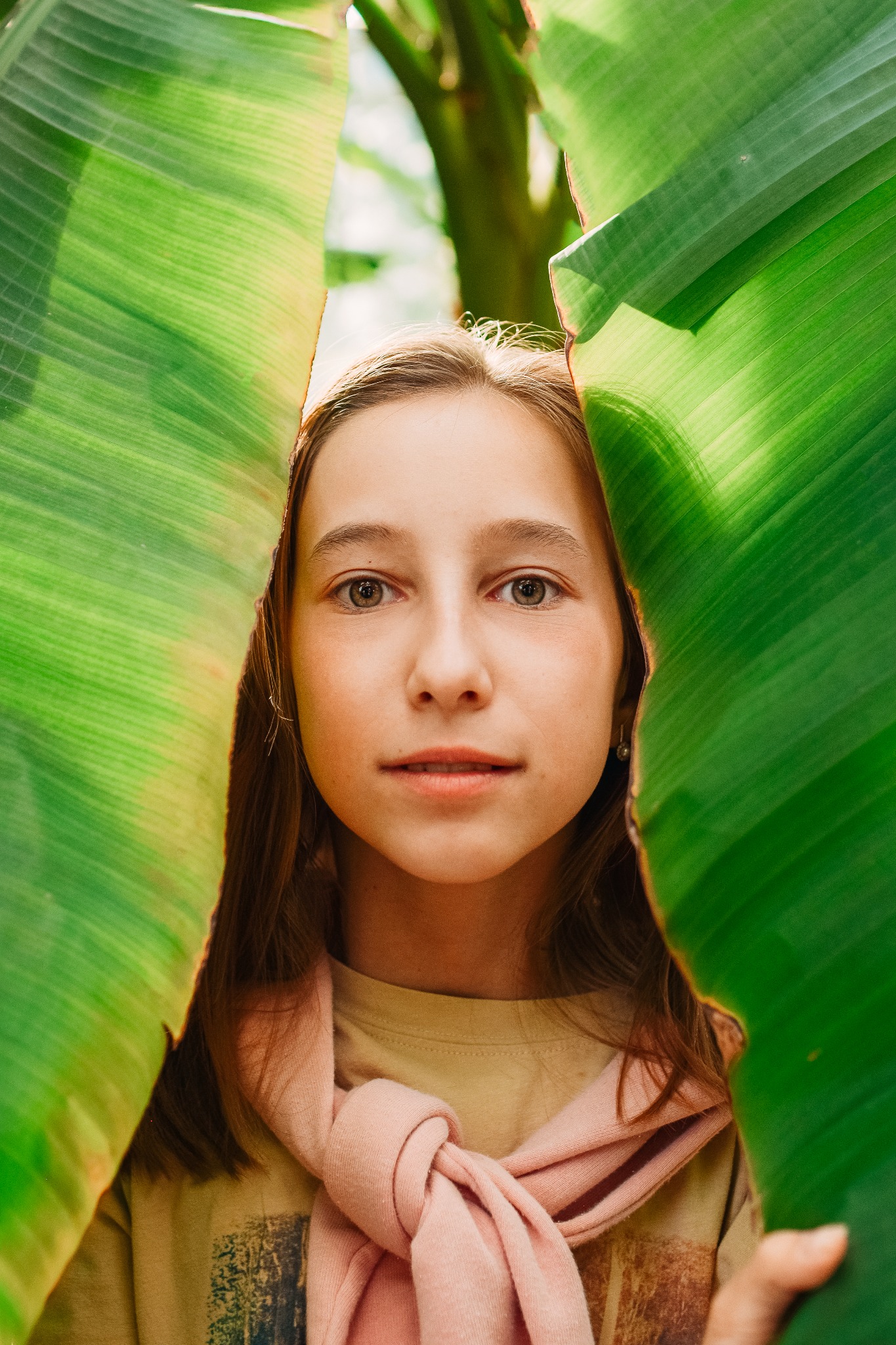 Portret młodej kobiety spoglądającej przez rozszczep w dużych, zielonych liściach bananowca, z delikatnym różowym szalem zawiązanym na szyi.