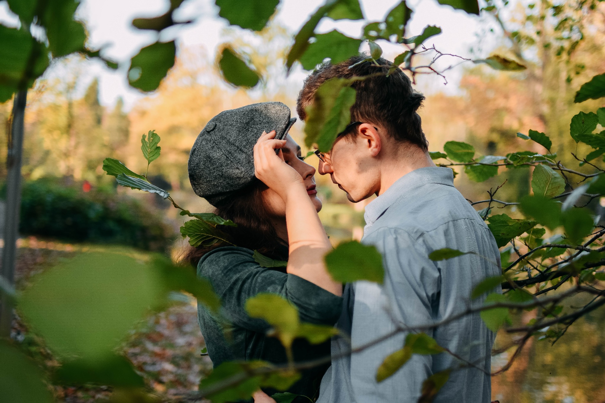 Para w parku, kobieta w kaszkiecie dotyka twarzy, czułe zbliżenie przez liście drzew, jesienne tło.