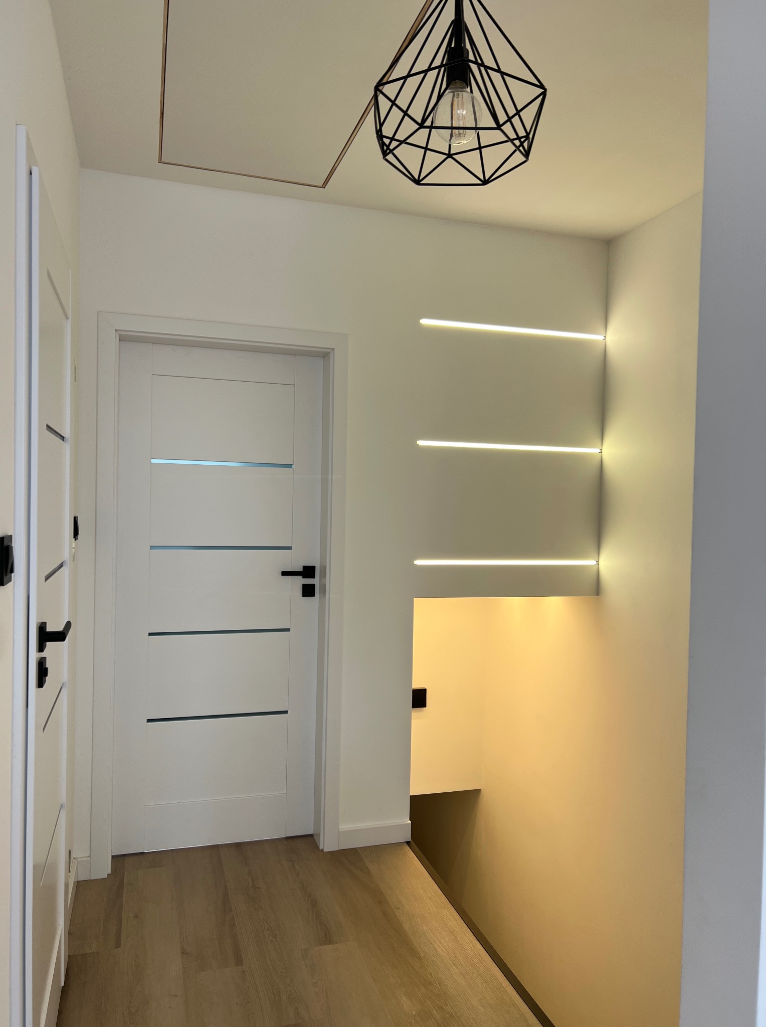 Nowoczesny przedpokój z białymi drzwiami, geometryczną lampą i trzema poziomymi paskami oświetlenia LED w ścianie. Widoczny fragment schodów.
