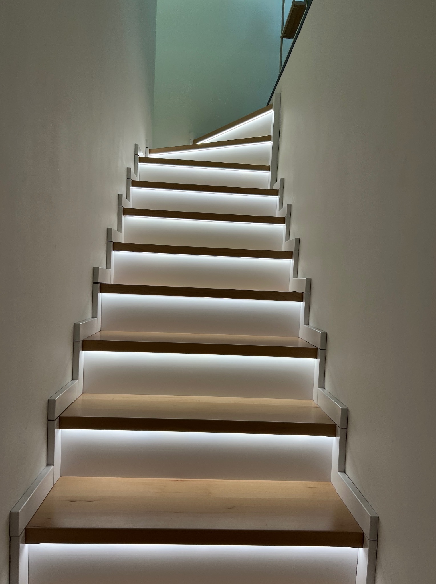 Nowoczesne schody z podświetleniem LED, drewniane stopnie z białymi listwami, oświetlenie wbudowane pod każdym stopniem, minimalistyczny design, jasne ściany.