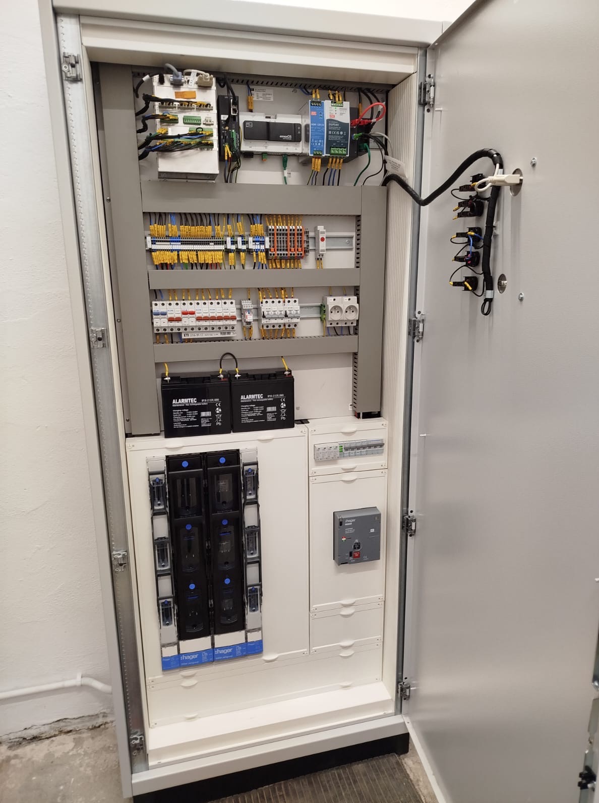 Otwarta szafa sterownicza z widocznymi elementami instalacji elektrycznej: bezpieczniki, przekaźniki, listwy zaciskowe i akumulatory. Szara obudowa, białe wnętrze.