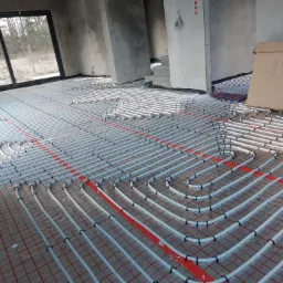 Szare rury ogrzewania podłogowego ułożone w pętle na czerwonej siatce w surowym wnętrzu, z kartonem z rysunkami rur w tle.