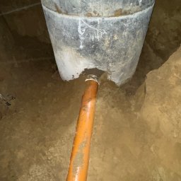 INSTAL-BUD KASZUBY SPÓŁKA Z OGRANICZONĄ ODPOWIEDZIALNOŚCIĄ - Szara studnia betonowa z podłączoną pomarańczową rurą kanalizacyjną w wykopie ziemnym. Widoczne ślady zaprawy i ziemi na elementach.