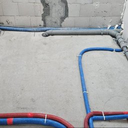 INSTAL-BUD KASZUBY SPÓŁKA Z OGRANICZONĄ ODPOWIEDZIALNOŚCIĄ - Ułożone rury ciepłej i zimnej wody (czerwone, niebieskie) oraz szare rury odpływowe na betonowej posadzce, z widocznymi punktami poboru wody i izolacją piankową w Starej Hucie.