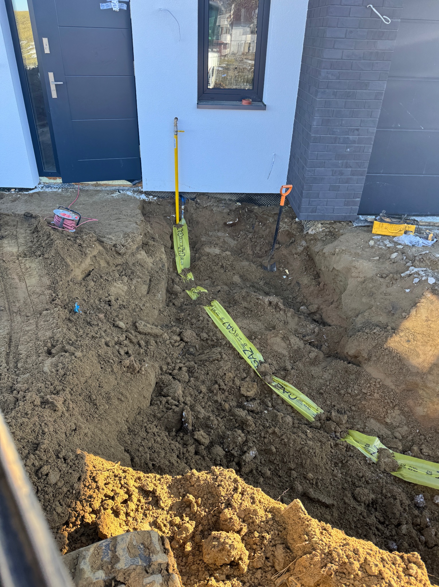 Wykop z żółtą taśmą ostrzegawczą 'GAZ' przy ścianie budynku z szarą stolarką okienną i drzwiami. Widoczne narzędzia: łopata i rura. Etap prac ziemnych.