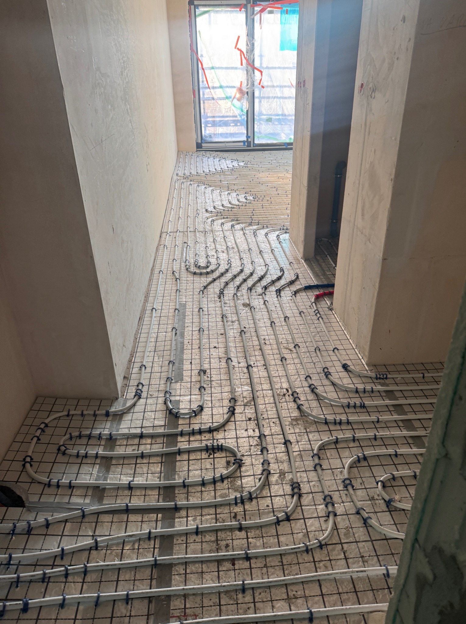 Ułożone rury ogrzewania podłogowego na siatce w surowym wnętrzu, widok z góry, przygotowane do zalania posadzką. Widoczne fragmenty ścian i okno w tle.